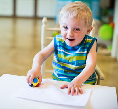 Interdisziplinäre Frühförderung. Bild zeigt Kind, dass auf ein Blatt Papier malt und in die Kamera schaut.