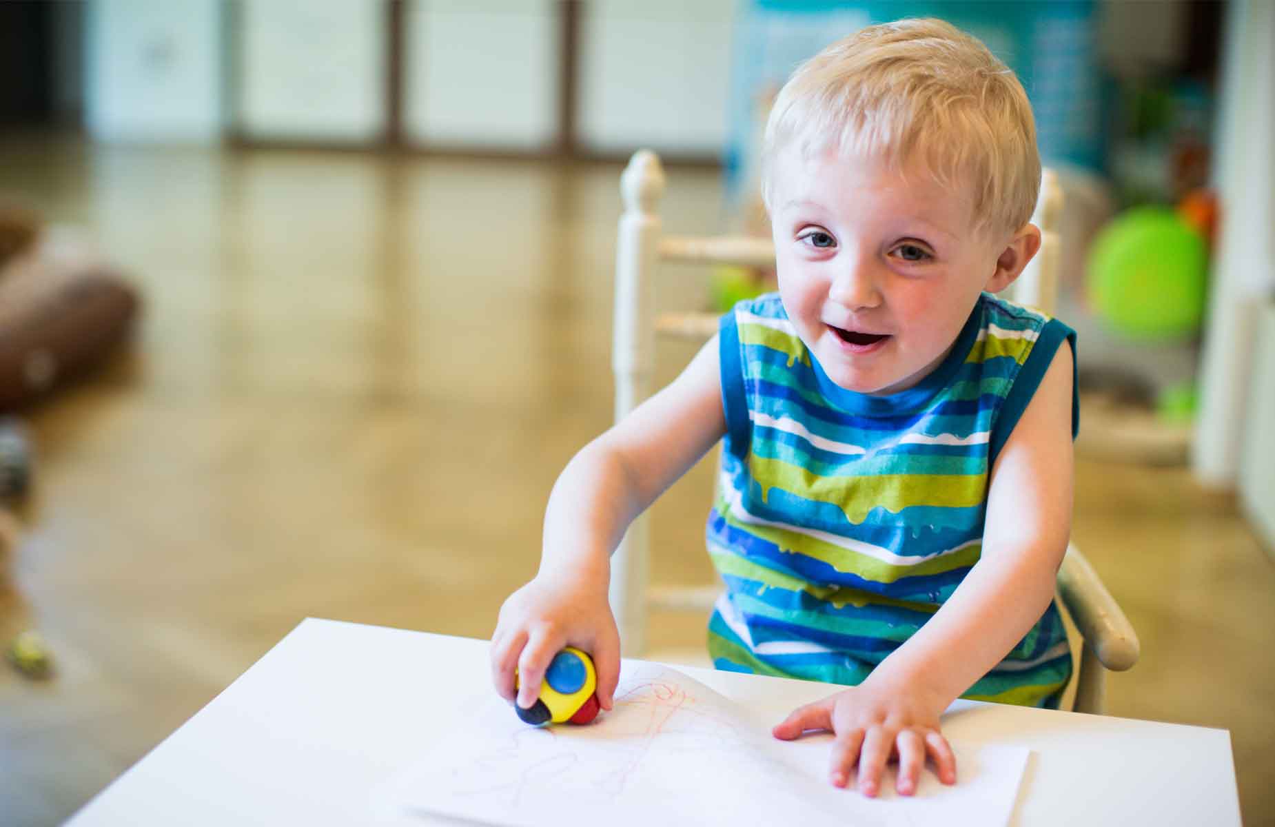 Interdisziplinäre Früförderung orientiert sich nach den Fähigkeiten des Kindes. Foto zeigt blonden Jungen an einem Tisch sitzend. Er malt mit einer besonderen Malkugel auf ein großes Blatt Papier.