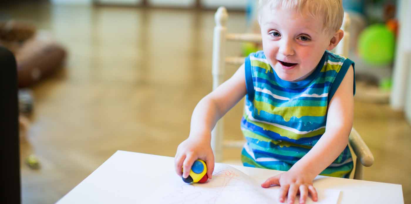 Interdisziplinäre Frühförderung. Bild zeigt Kind, dass auf ein Blatt Papier malt und in die Kamera schaut.